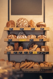 In den Regalen einer Bäckerei sind verschiedene handwerklich hergestellte Brote und Brötchen ausgestellt, mit Preisschildern unter jeder Sorte. Die Brote unterscheiden sich in Form, Größe und Krustentextur. Ein leeres Schild hängt über den Regalen.