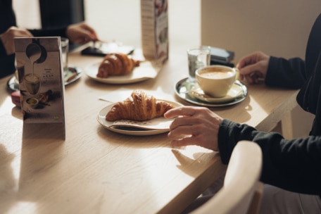 Zwei Personen sitzen an einem sonnenbeschienenen Tisch mit Croissants auf Tellern, Cappuccino in einer Tasse, Gläsern mit Wasser und Speisekarten in der Nähe. Eine Hand greift nach einem Croissant, die andere hält ein Telefon.