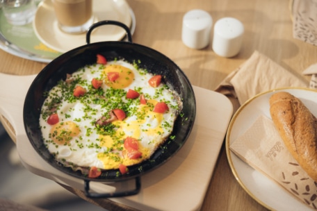 Eine Pfanne mit Spiegeleiern, die mit Schnittlauch und Tomatenwürfeln belegt sind, steht auf einem Holzbrett neben einem Teller mit einem Brötchen, im Hintergrund stehen Salz- und Pfefferstreuer und ein Kaffee.
