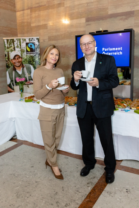 Eine Frau und ein Mann, beide in Geschäftskleidung, stehen mit Kaffeetassen in der Hand lächelnd vor einem Buffettisch. Hinter ihnen sind ein Fairtrade-Banner und ein Monitor mit der Aufschrift "Parlament Österreich" zu sehen.