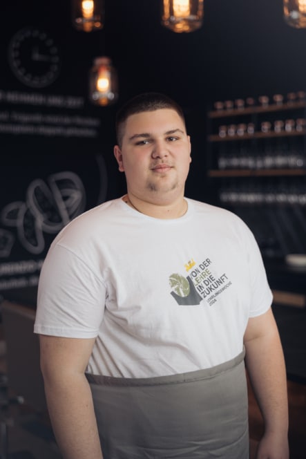 Ein junger Mann mit kurzen dunklen Haaren, der ein weißes T-Shirt mit einer Grafik und einem Text sowie eine graue Schürze trägt, steht in einem Raum mit Hängelampen und einer Kreidetafel im Hintergrund.