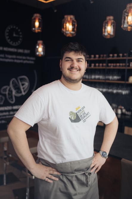 Ein lächelnder Mann mit kurzen dunklen Haaren steht in einem modernen Café, trägt ein weißes T-Shirt und eine graue Schürze, die Hände in die Hüften gestemmt. Im Hintergrund sind warme Hängelampen und Tafeln mit Schrift zu sehen.