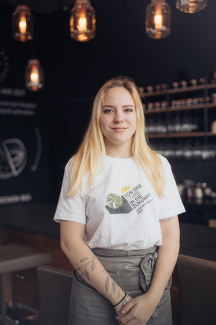 Eine junge Frau mit langen blonden Haaren steht lächelnd in einem Haus, trägt ein weißes T-Shirt und eine graue Schürze. Im Hintergrund sind Hängelampen, Regale mit Gläsern und eine dunkle Wand mit Schrift zu sehen.