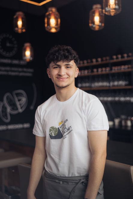 Ein junger Mann mit lockigem Haar lächelt, während er in einem stilvollen, modernen Café steht. Er trägt ein weißes T-Shirt mit Logo und Text und eine Schürze. Im Hintergrund sind Regale und warme Hängelampen zu sehen.