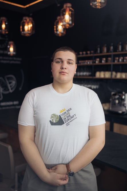 Eine junge Person mit kurzen Haaren steht in einem Innenraum und trägt ein weißes T-Shirt mit einer Grafik und deutschem Text. Im sanft beleuchteten Hintergrund sind warme Hängelampen und Regale mit Gläsern und Kaffeetassen zu sehen.