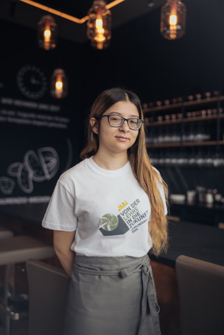 Eine junge Frau mit langen braunen Haaren und Brille steht in einem modernen Café. Sie trägt ein weißes T-Shirt mit einer grünen und gelben Grafik und eine graue Schürze. Im Hintergrund sind warme Hängelampen und Regale mit Gläsern zu sehen.