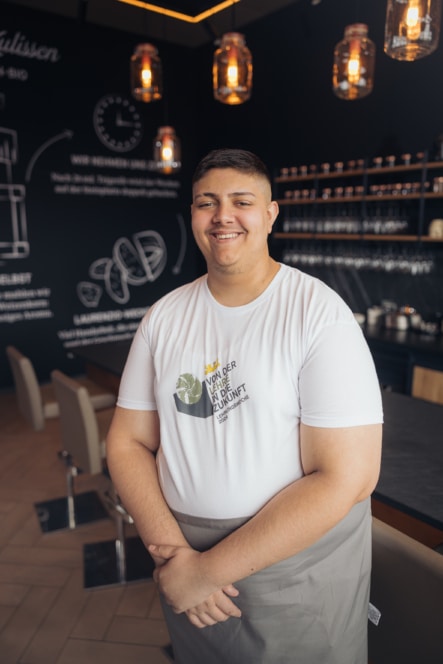 Eine lächelnde Person mit weißem T-Shirt und grauer Schürze steht in einem modernen Café mit dunklen Wänden, Hängelampen und Regalen mit Glaswaren im Hintergrund.