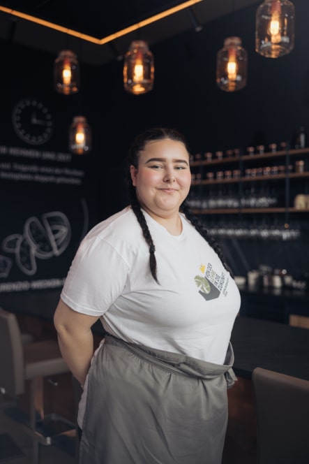 Eine Frau mit geflochtenen Haaren steht lächelnd hinter einem Bartresen in einem modernen Café und trägt ein weißes T-Shirt und eine graue Schürze. Im Hintergrund sind warme Hängelampen und Regale mit Gläsern zu sehen.