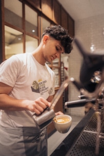 Ein junger Barista mit lockigem Haar gießt gedämpfte Milch in eine Tasse Kaffee und kreiert so einen Milchkaffee, während er hinter dem Tresen eines Cafés steht. Er trägt ein weißes T-Shirt und eine Schürze.