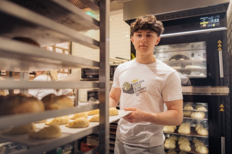 Ein junger Mann in einem weißen Hemd hält ein Tablett mit ungebackenen Brötchen in einer Bäckereiküche und steht zwischen Brotständern in der Nähe eines Industrieofens.