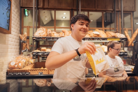 Eine lächelnde Bäckereimitarbeiterin reicht einem Kunden eine Papiertüte über den Tresen, während im Hintergrund Brot und Gebäck in Regalen ausgestellt sind. Eine andere Angestellte steht in der Nähe an der Kasse.