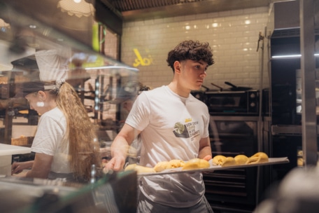Ein junger Mann in einem weißen T-Shirt hält ein Tablett mit ungebackenem Gebäck in einer Großküche, während eine andere Person in der Nähe arbeitet. Im Hintergrund sind Öfen und Küchengeräte zu sehen.