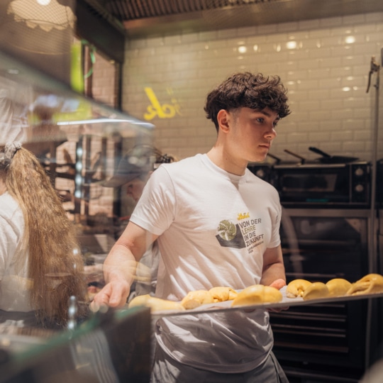 Ein junger Mann in einem weißen T-Shirt hält ein Tablett mit ungebackenem Gebäck in einer Großküche, während eine andere Person in der Nähe arbeitet. Im Hintergrund sind Öfen und Küchengeräte zu sehen.