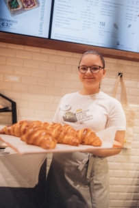 Eine Frau mit Brille und Schürze lächelt, während sie ein Tablett mit frisch gebackenen Croissants in einer Bäckerei hält. An der Wand hinter ihr ist eine Speisekarte zu sehen.