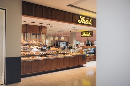 In einer Bäckerei in einem Einkaufszentrum werden hinter Glas verschiedene Backwaren und Brot angeboten. Warme Beleuchtung, eine Ströck-Leuchtreklame und Holzdekor schaffen eine gemütliche Atmosphäre. Das Personal ist hinter der Theke zu sehen.