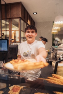 Eine lächelnde Bäckereimitarbeiterin im weißen Hemd steht hinter einer Theke und streckt der Kamera ein Gebäckstück entgegen. Im Hintergrund sind Auslagen, Regale und ein weiterer Mitarbeiter zu sehen.