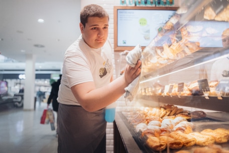 Ein junger Mann mit weißem T-Shirt und grauer Schürze reinigt die Glasvitrine einer Konditorei in einer hell erleuchteten Bäckerei, in der verschiedene Backwaren zu sehen sind.