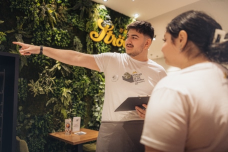 Zwei Angestellte in weißen Hemden stehen in einem Restaurant mit einer grünen Pflanzenwand; der eine hält ein Klemmbrett in der Hand und zeigt auf den anderen, der aufmerksam zuhört.