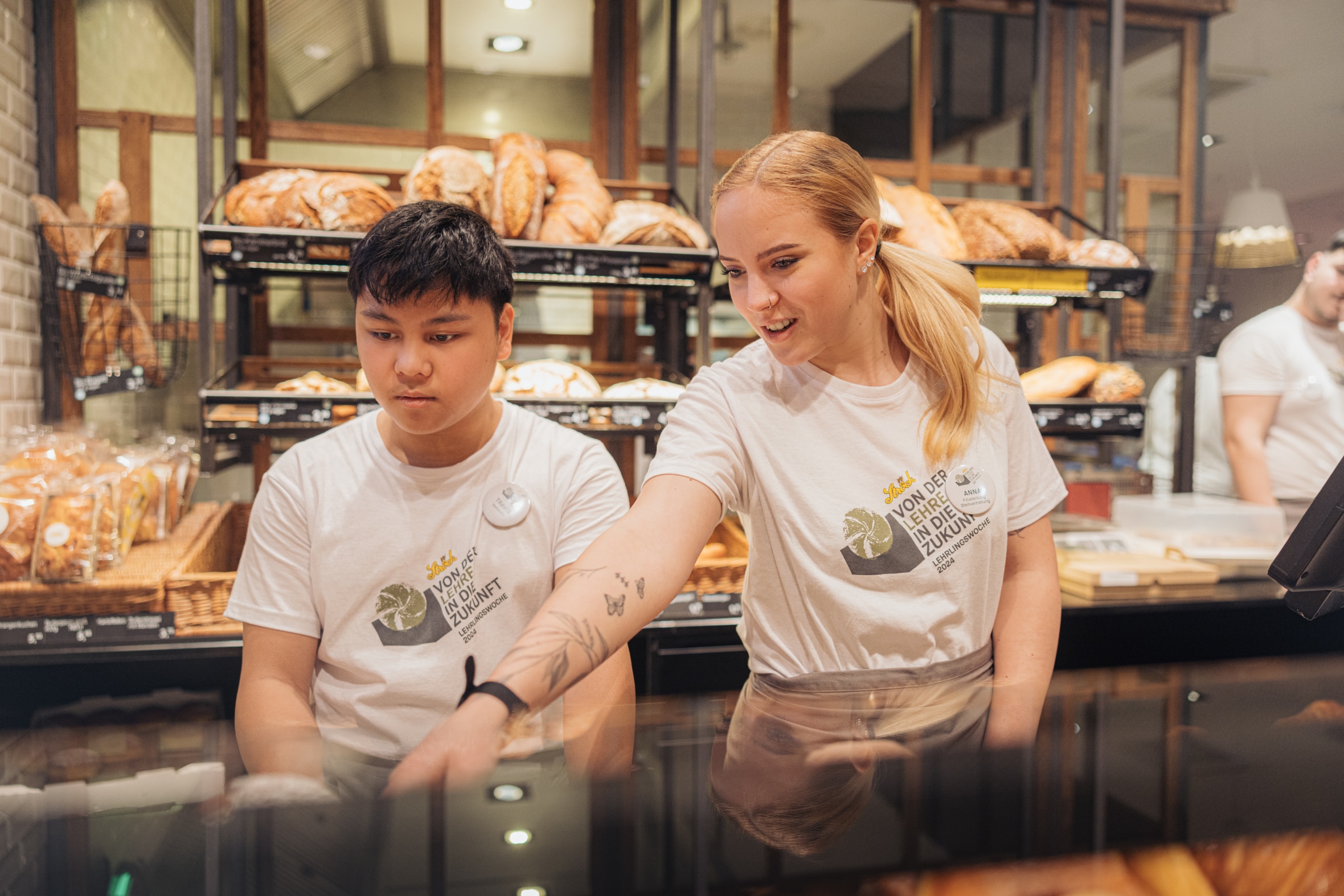 Zwei Bäckereiangestellte in weißen Hemden stehen hinter einer Theke, hinter der sich Regale mit Brot befinden. Einer zeigt auf etwas auf der Theke, während der andere aufmerksam zuschaut.
