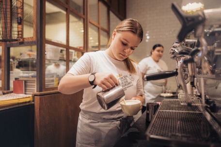 Ein Barista in weißem Hemd und Schürze gießt an der Theke eines Coffeeshops dampfende Milch in eine Tasse, während ein anderer Angestellter unscharf im Hintergrund neben einer Espressomaschine zu sehen ist.