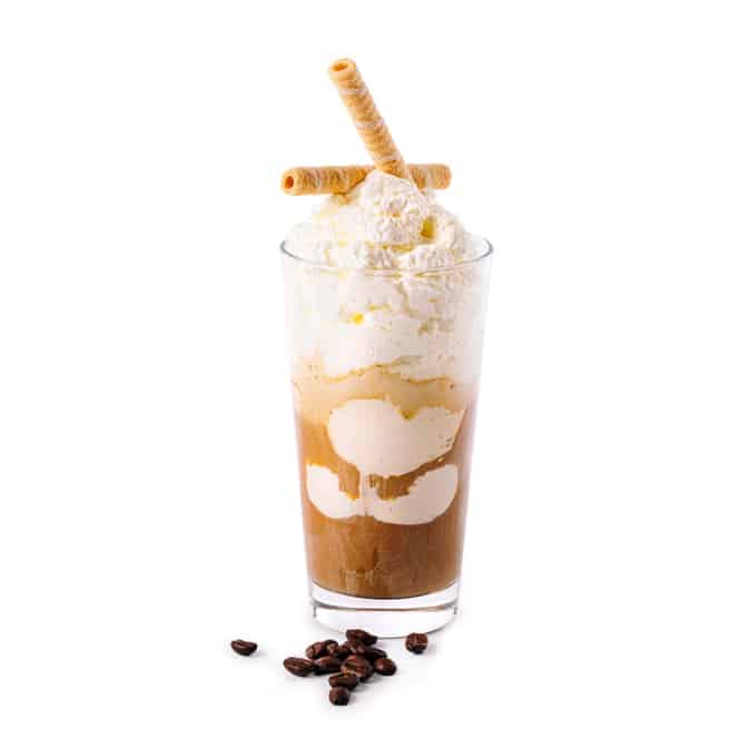 Ein Glas Eiskaffee mit Schlagsahne und zwei Waffelröllchen, auf deren Boden einige Kaffeebohnen verstreut sind, alles vor einem weißen Hintergrund.