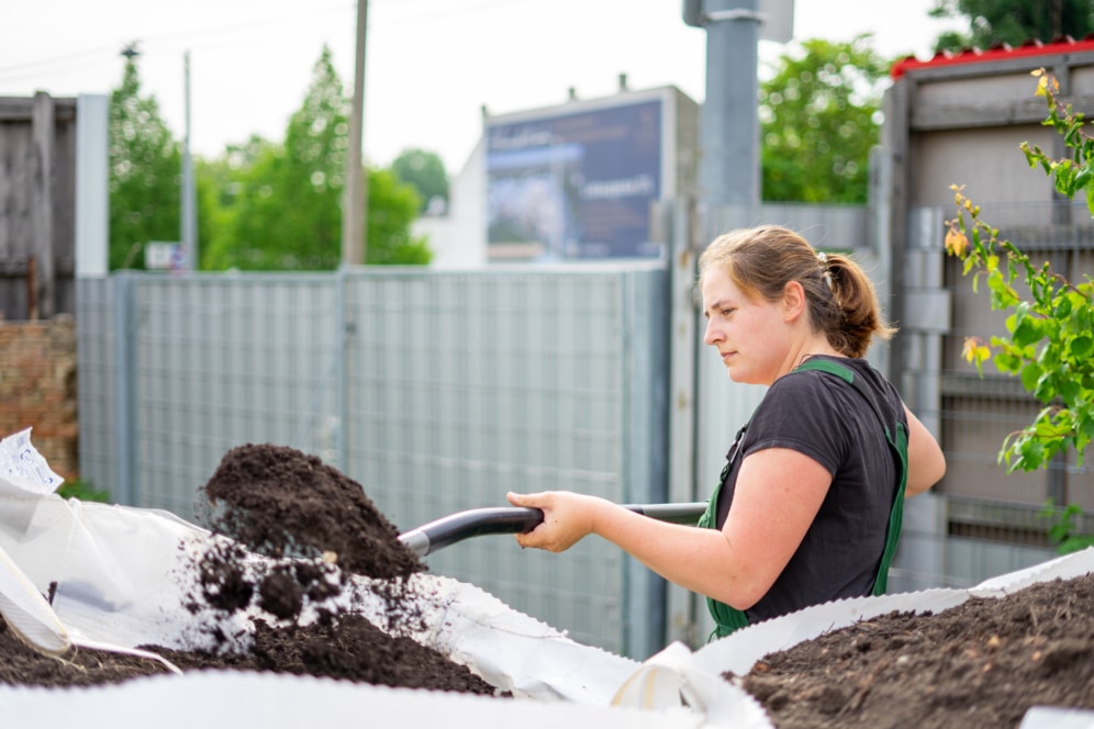 Eine Frau in Arbeitskleidung bewegt mit einer Schaufel Erde oder Kompost im Freien, mit einem Metallzaun und Grünpflanzen im Hintergrund.