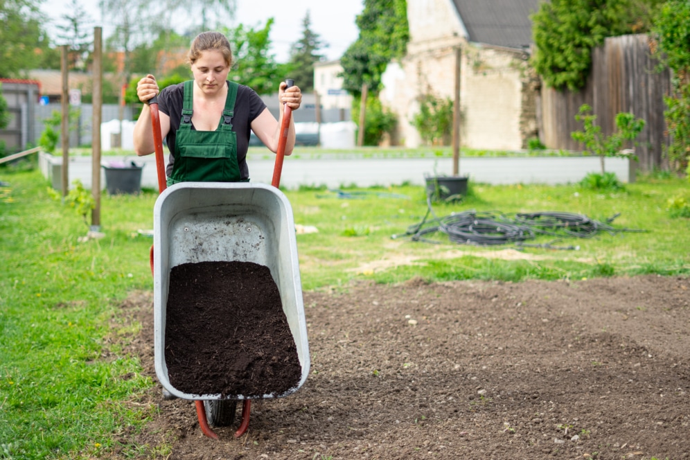 Eine Person in grünem Overall kippt eine mit Erde gefüllte Schubkarre auf ein Gartenbeet in einer Gartenanlage im Freien. Im Hintergrund sind grünes Gras und Gartenbeete zu sehen.