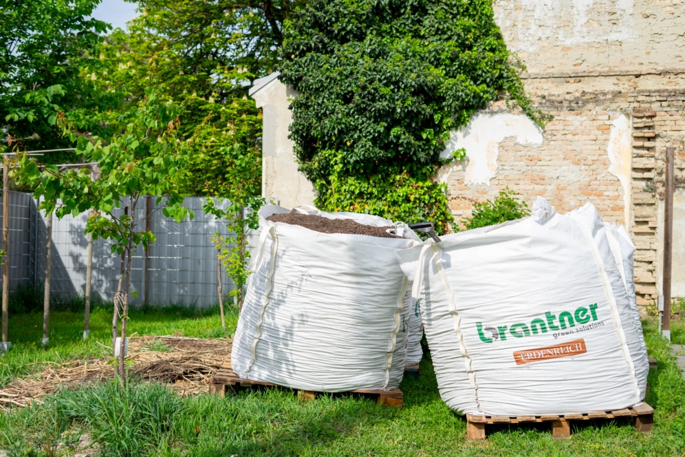 Zwei große weiße Säcke, die mit Erde oder Kompost gefüllt sind, stehen auf Holzpaletten in einem grünen Garten, mit einem hohen Baum, Pflanzen und einer Ziegelmauer im Hintergrund. Der vordere Sack ist mit "Lorantner" beschriftet.
