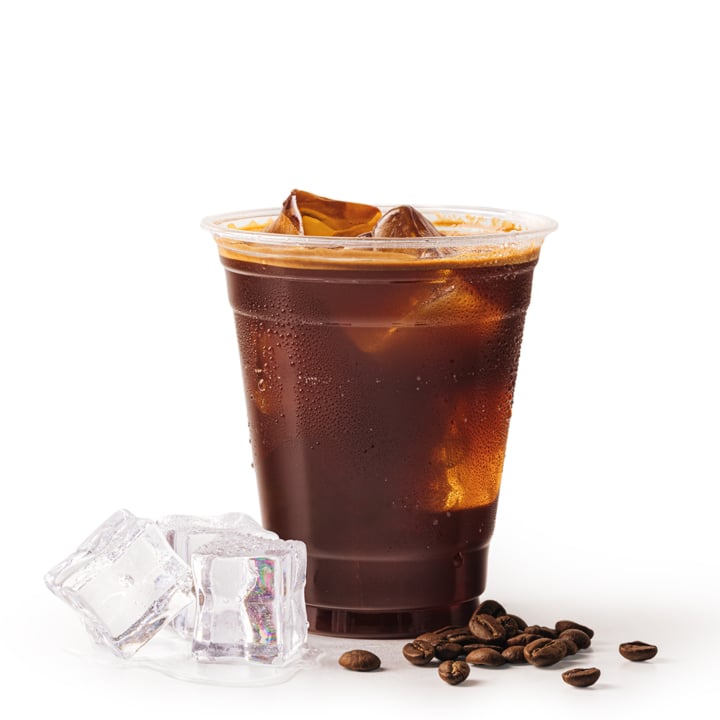 Ein mit schwarzem Eiskaffee gefüllter Plastikbecher, der mit Eiswürfeln gekrönt ist. Neben dem Becher befinden sich einige lose Kaffeebohnen und mehrere Eiswürfel auf weißem Hintergrund.