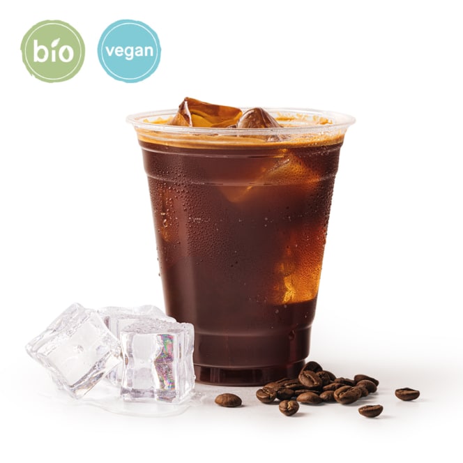 Ein durchsichtiger Plastikbecher, gefüllt mit Eiskaffee, steht neben Eiswürfeln und Kaffeebohnen. Zwei runde Etiketten über dem Becher tragen die Aufschrift Bio und Vegan. Der Hintergrund ist weiß.