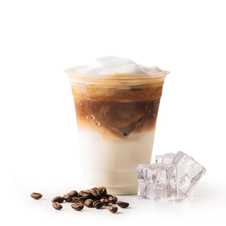 Ein Plastikbecher mit eisgekühltem Milchkaffee, Milch und Espresso, gekrönt von Schaum, steht neben Eiswürfeln und Kaffeebohnen auf einem weißen Hintergrund.