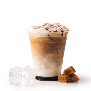 Ein Plastikbecher, gefüllt mit eisgekühltem Karamell-Macchiato, gekrönt von Schaum und Karamellspritzern. Neben dem Becher sind zwei Eiswürfel und drei Karamellbonbons zu sehen, alles vor einem weißen Hintergrund.