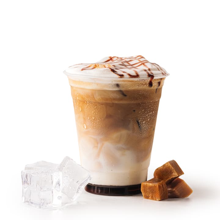 Ein Plastikbecher, gefüllt mit eisgekühltem Karamell-Macchiato, gekrönt von Schaum und Karamellspritzern. Neben dem Becher sind zwei Eiswürfel und drei Karamellbonbons zu sehen, alles vor einem weißen Hintergrund.