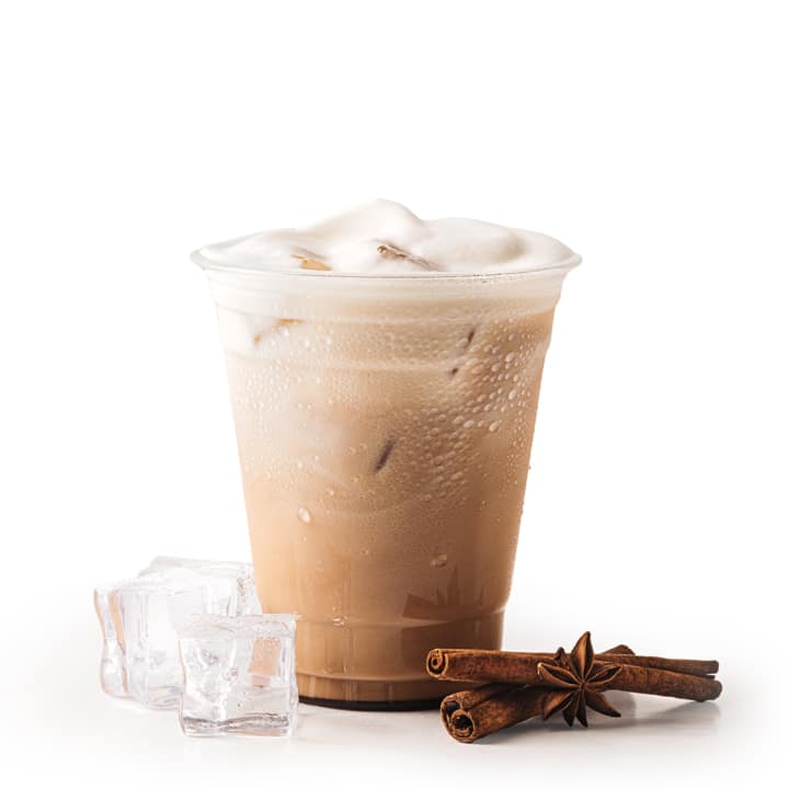 Ein Plastikbecher mit eisgekühltem Milchkaffee und Schaum darauf, daneben drei Eiswürfel und ein Bündel Zimtstangen mit Sternanis, alles vor einem weißen Hintergrund.