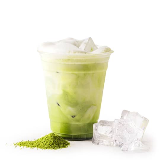 Ein Plastikbecher, gefüllt mit einem eisgekühlten grünen Matcha-Latte, mit Schaum bedeckt, steht neben drei Eiswürfeln und einem kleinen Haufen grünem Matcha-Pulver auf einem weißen Hintergrund.