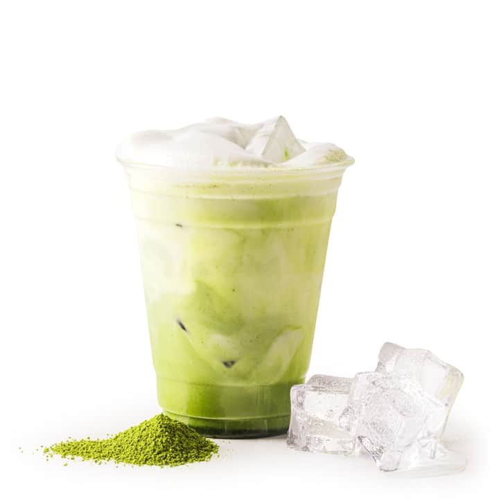 Ein Plastikbecher, gefüllt mit einem eisgekühlten grünen Matcha-Latte, mit Schaum bedeckt, steht neben drei Eiswürfeln und einem kleinen Haufen grünem Matcha-Pulver auf einem weißen Hintergrund.