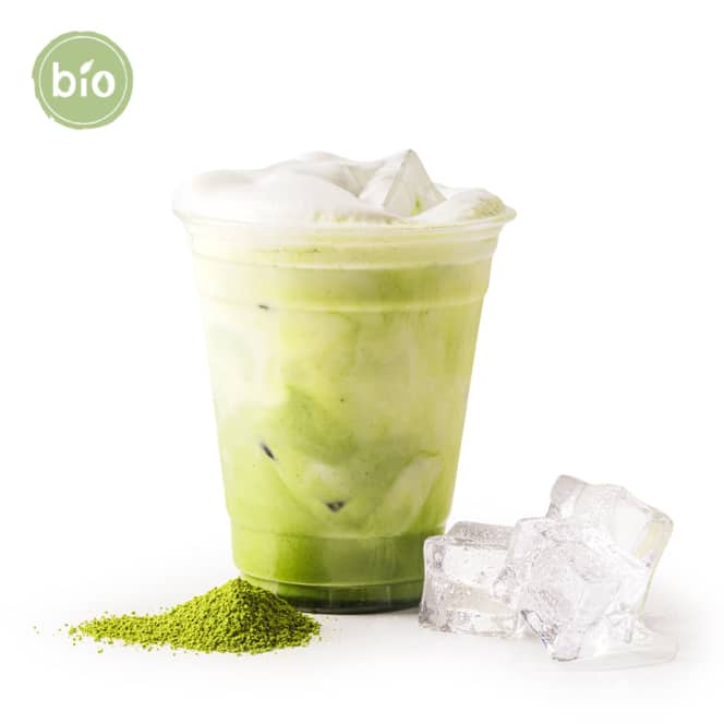 Ein Plastikbecher mit eisgekühltem Matcha-Latte mit Schaumkrone steht neben Eiswürfeln und einem Haufen Matcha-Pulver. In der oberen linken Ecke befindet sich ein grünes Bio-Etikett, das anzeigt, dass das Getränk aus biologischem Anbau stammt. Der Hintergrund ist weiß.