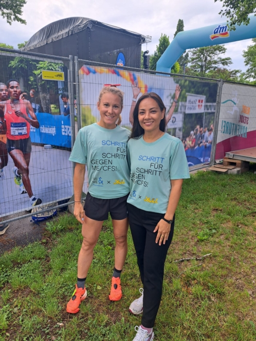 Zwei Frauen stehen nebeneinander im Gras und lächeln in die Kamera. Sie tragen hellblaue Hemden mit deutscher Aufschrift und Sportschuhe. Im Hintergrund sind ein Laufbanner und eine Veranstaltungsanordnung zu sehen.