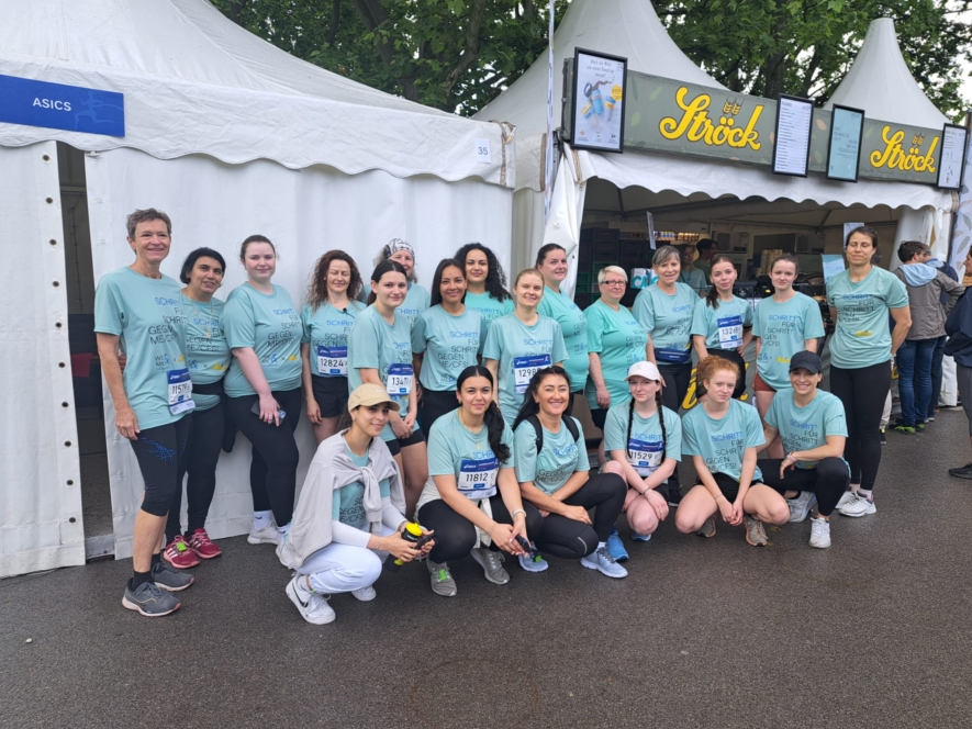 Eine Gruppe von Frauen in passenden hellblauen Hemden und Laufklamotten posiert gemeinsam vor weißen Zelten bei einer Laufveranstaltung und lächelt in die Kamera. Einige tragen Startnummern und Mützen, und der Boden scheint nass vom Regen.
