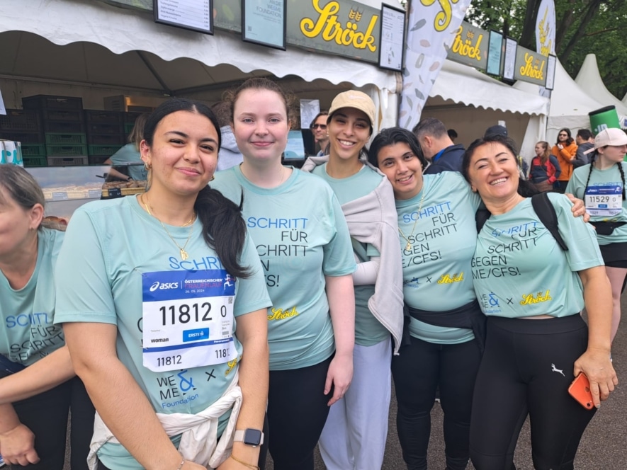 Fünf Frauen in passenden hellblauen Schritt für Schritt gegen ME/CFS-Shirts lächeln und posieren gemeinsam bei einer Veranstaltung im Freien, während im Hintergrund Essensstände und andere Teilnehmer zu sehen sind.