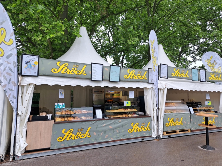 Bäckereistand im Freien mit grünem "Ströck"-Schild, der Gebäck und Brot unter einem weißen Vordach anbietet. Darüber sind Speisekarten angebracht, und im Hintergrund sind Bäume zu sehen. Die Szene scheint auf einem Markt oder einem Fest zu spielen.