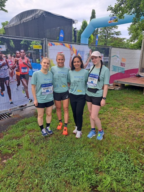 Vier Frauen in Laufklamotten posieren gemeinsam bei einer Laufveranstaltung im Freien. Sie tragen passende grüne Hemden und Startnummern und stehen auf einer Wiese mit einer Bühne, Veranstaltungsbannern und einem großen aufblasbaren Bogen im Hintergrund.