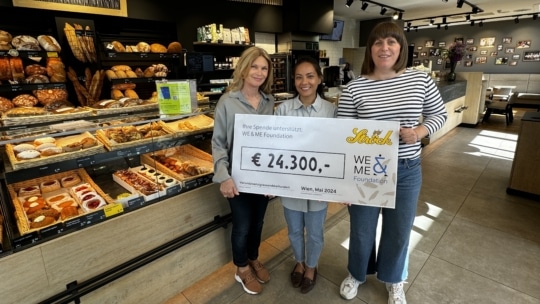 Drei Frauen stehen lächelnd in einer Bäckerei und halten einen großen Spendenscheck über 24.300 € für die WE & ME Foundation in der Hand. Hinter ihnen sind Regale mit Brot und Gebäck zu sehen.