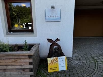 Ein Schild mit der Aufschrift Fuchssteiner Bio Imkerei ist an einer weißen Wand über einer braunen Bienenskulptur aus Metall angebracht. Davor steht ein gelbes Schild mit der Aufschrift Bio-Imker/in des Jahres und einer Wabengrafik. Auf der linken Seite steht ein Pflanzgefäß.