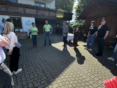 Eine Gruppe von Menschen steht an einem sonnigen Tag in einem Innenhof, einige hören zwei Personen zu, die in der Mitte sprechen. Schatten erstrecken sich über das Kopfsteinpflaster, und im Hintergrund ist ein Holzgebäude zu sehen.