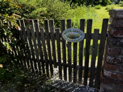 Ein hölzernes Gartentor mit einem Schild mit der Aufschrift "Natur im Garten" in deutscher Sprache, umgeben von Grün und teilweise von Sonnenlicht beschattet. Neben dem Tor sind ein Steinweg und eine Ziegelmauer zu sehen.