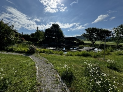Ein Kiesweg schlängelt sich durch einen grasbewachsenen Garten voller Wildblumen zu einem Teich, der von Bäumen umgeben ist, unter einem sonnigen, teilweise bewölkten Himmel. Im Hintergrund sind ein Holzgebäude und in der Ferne grüne Hügel zu sehen.