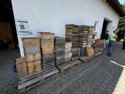 Stapel von Holzkisten und Paletten stapeln sich vor einer weißen Gebäudewand. Ein paar Leute stehen in der Nähe auf einem sechseckigen gefliesten Weg. Ein grünes Bio-Zertifizierungsschild ist an der Wand zu sehen.