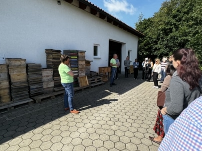 Eine Gruppe von Menschen steht vor einem weißen Gebäude mit gestapelten Holzkisten. Eine Frau in einem grünen Hemd spricht zu der Gruppe, während eine andere Person in der Nähe der offenen Tür des Gebäudes steht. Im Hintergrund sind Bäume und Sonnenlicht zu sehen.