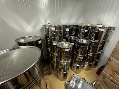 An den Wänden eines hell getäfelten Lagerraums stapeln sich glänzende Metallfässer, einige mit Etiketten und Hähnen. Auf einigen Fässern stehen Gläser, und auf dem Boden sind Holzpaletten zu sehen.